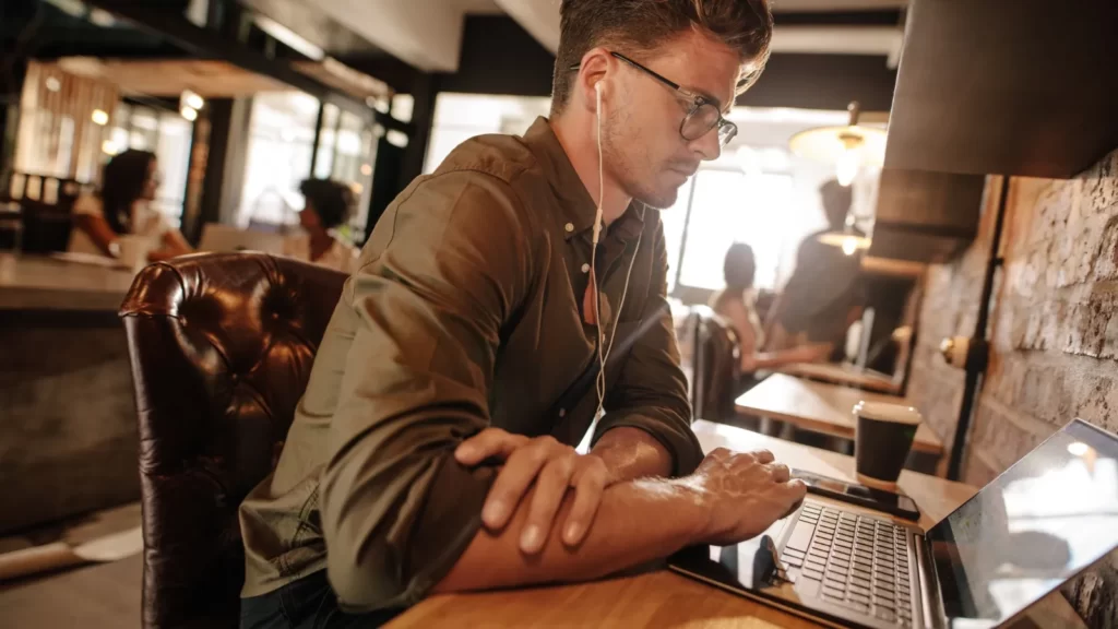 Man wearing in-ear headphones working with laptop in a café